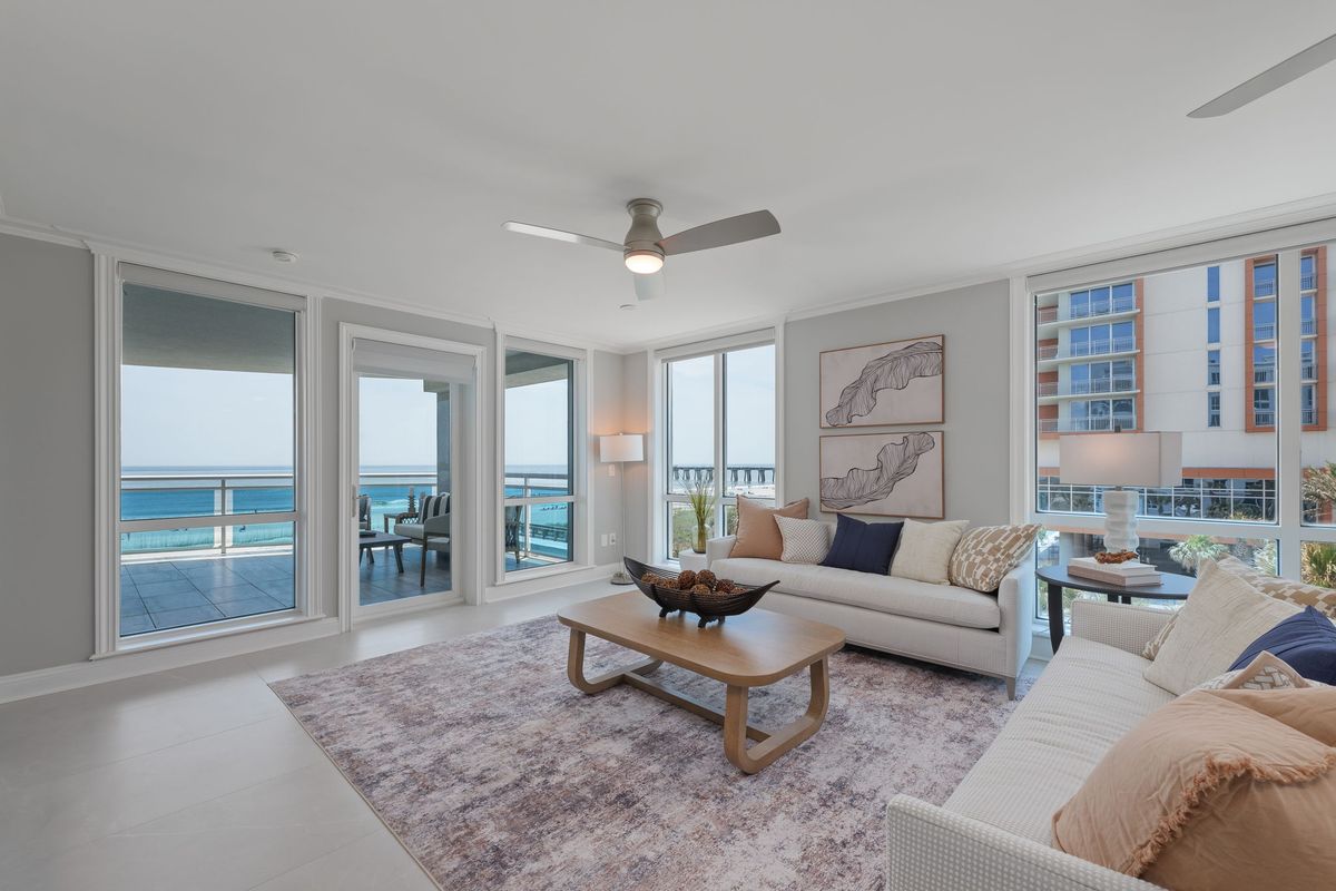 Living room with floor-to-ceiling windows and a balcony overlooking a turquoise ocean, with white sofas and a wood coffee table