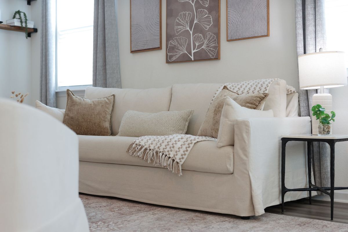 Cream slipcovered sofa with textured pillows and a knit throw, a table lamp and framed botanical art on the wall