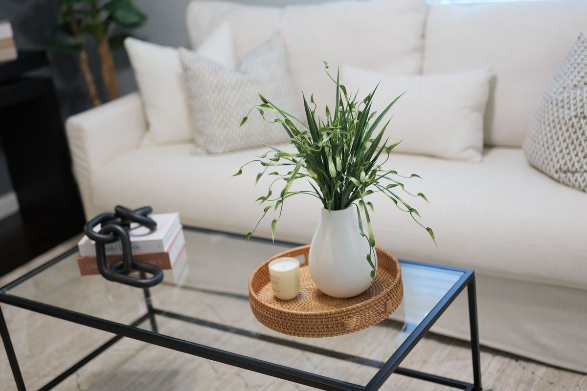 Glass coffee table styled with a white vase of greenery, a woven tray with a candle and stacked books, in front of an ivory sofa