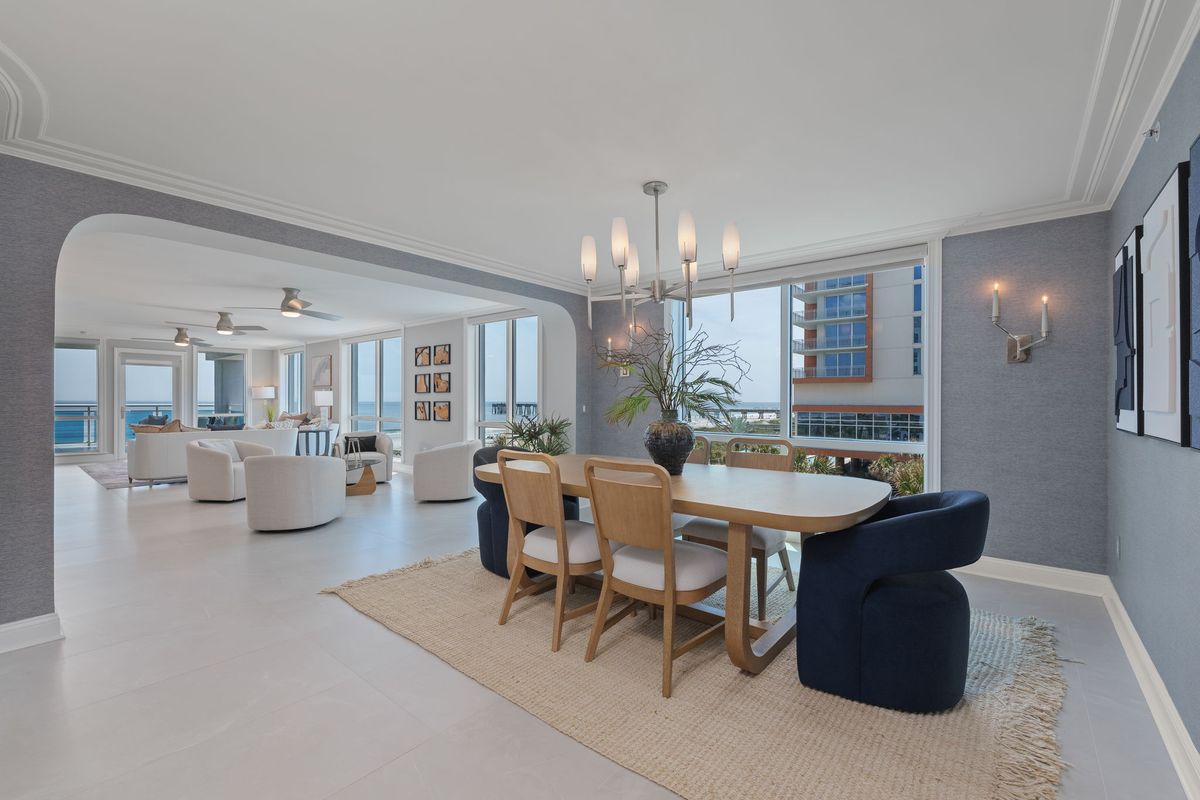 Dining table with navy upholstered chairs and a chandelier, with an archway to the living area and the ocean beyond